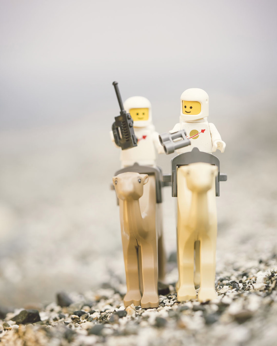 Two LEGO classic spacemen riding camels across a rocky beach. One is holding binoculars and one is holding a walkie talkie.