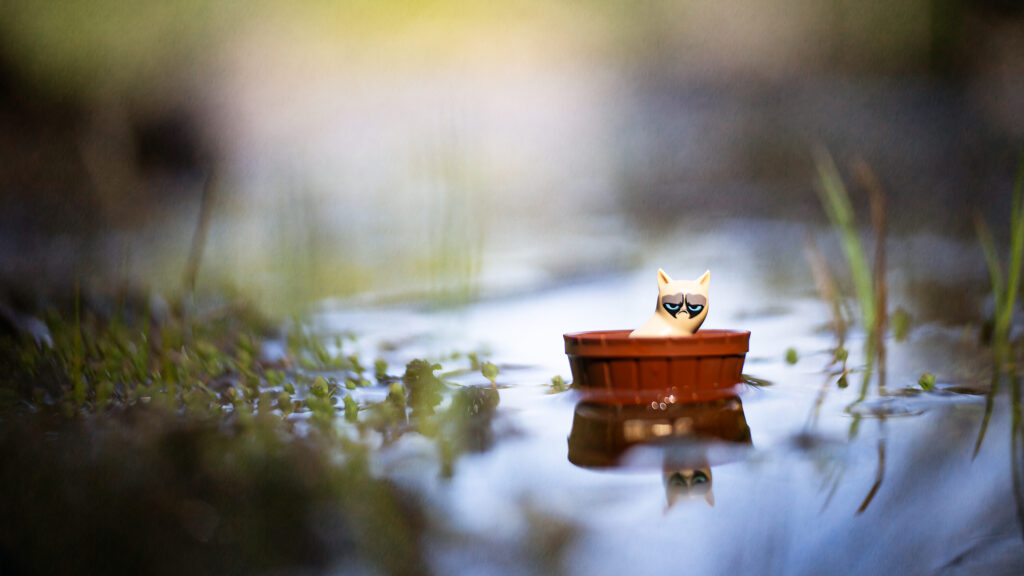 A toy cat with a grumpy expression sits in a small, brown, wooden barrel floating on calm water.