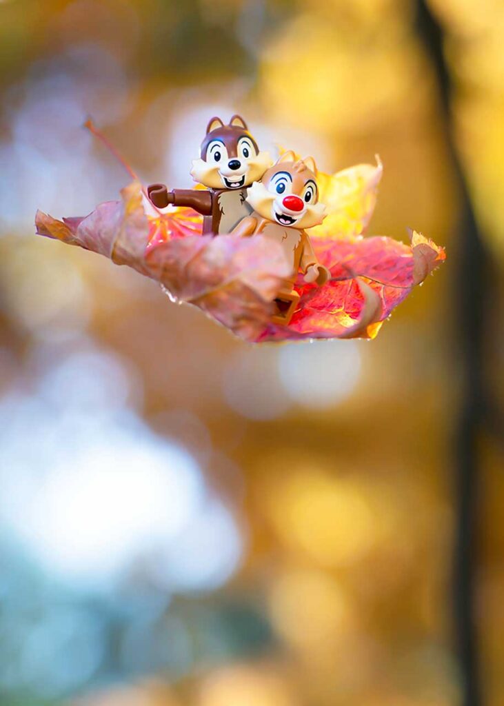 Two animated chipmunks riding on a colorful autumn leaf against a blurred backdrop of yellow and orange foliage.