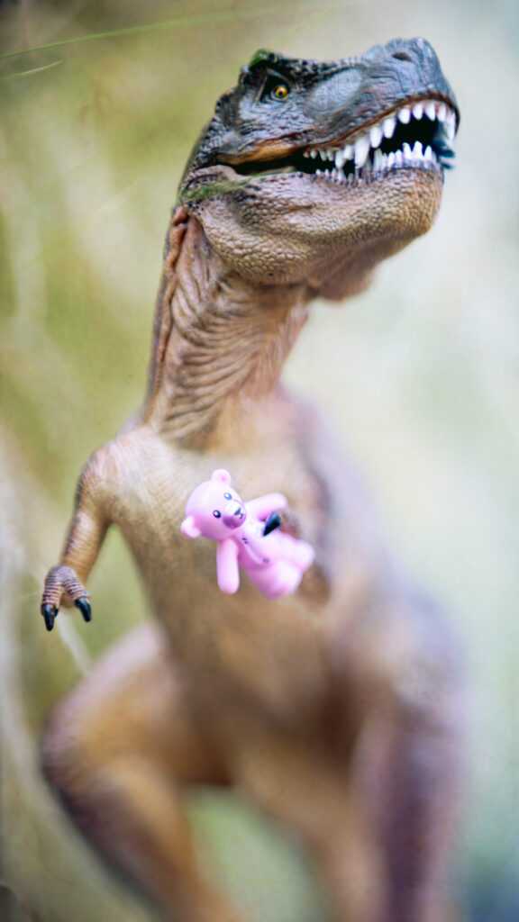 A realistic toy Tyrannosaurus Rex with its mouth open, displaying sharp teeth, clutches a small pink teddy bear in its clawed hand.