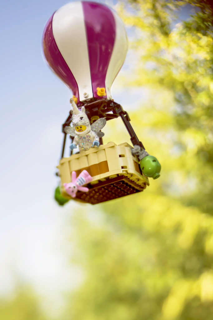 A Lego hot air balloon with a unicorn minifigure and a pink bunny in the basket floats against a blurred background of green foliage and blue sky. The pink bunny has fallen from the unicorns hands and a look of horror crosses her face.